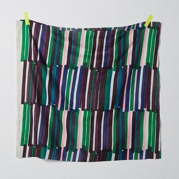 A yard of Nani IRO double gauze fabric hanging on a light gray wall. The fabric has slanted, irregular stripes, about 3cm wide and 30cm long. The stripes are cobalt blue, emeral green, teal, rose pink and shades of purple on a white background.